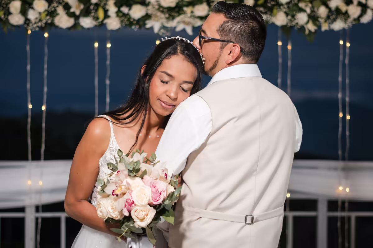 Novios abrazados con el ramo de novia frente a un telón de flores blancas