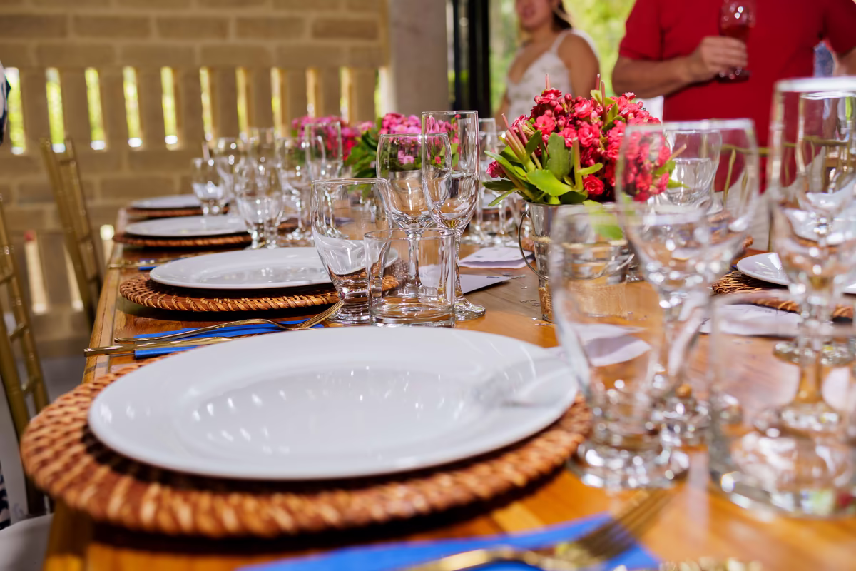 Mesa larga montada con bajoplatos tejidos, cubiertos dorados y flores fucsia