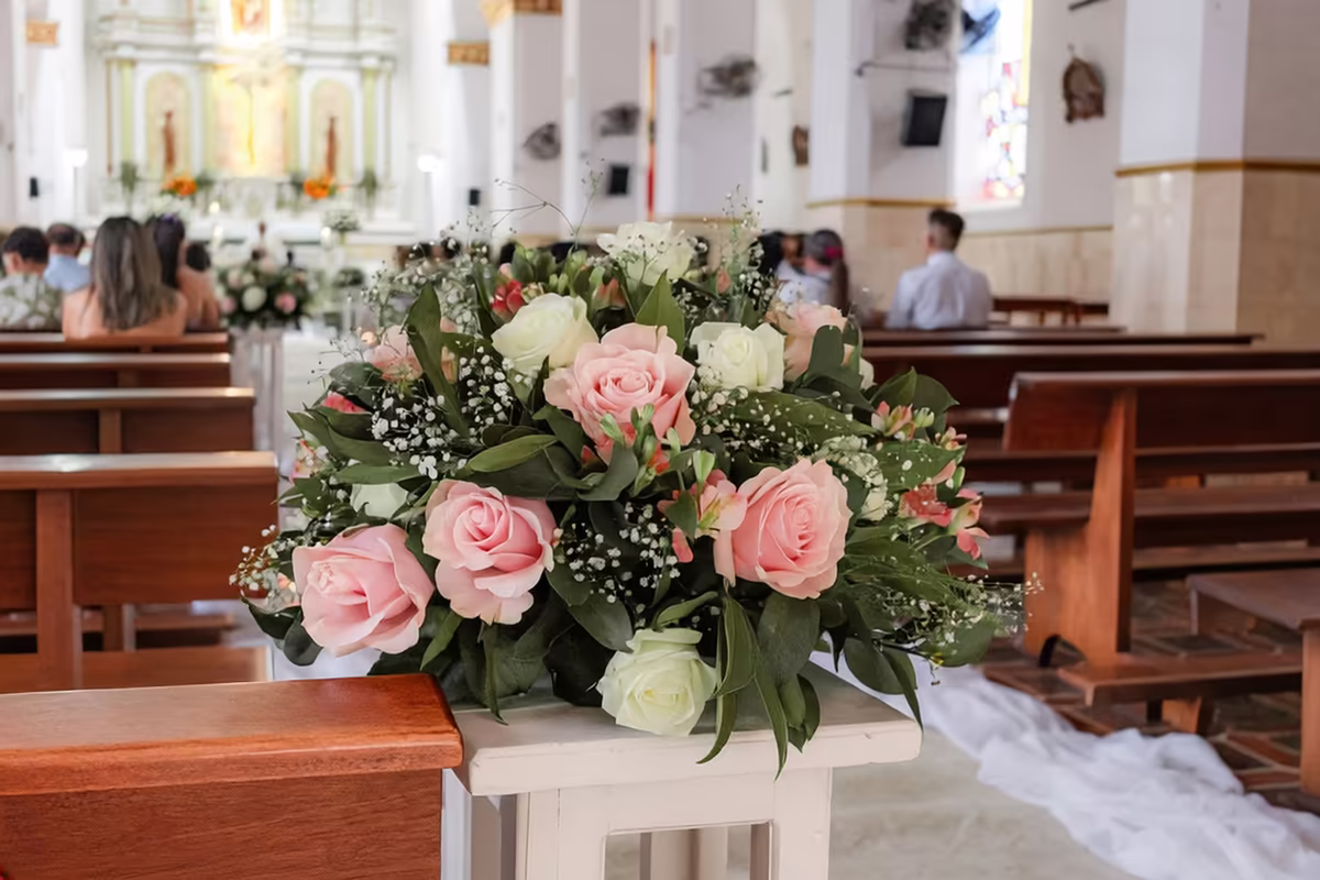 Arreglo de rosas rosadas y blancas sobre un pedestal junto a las bancas de la iglesia