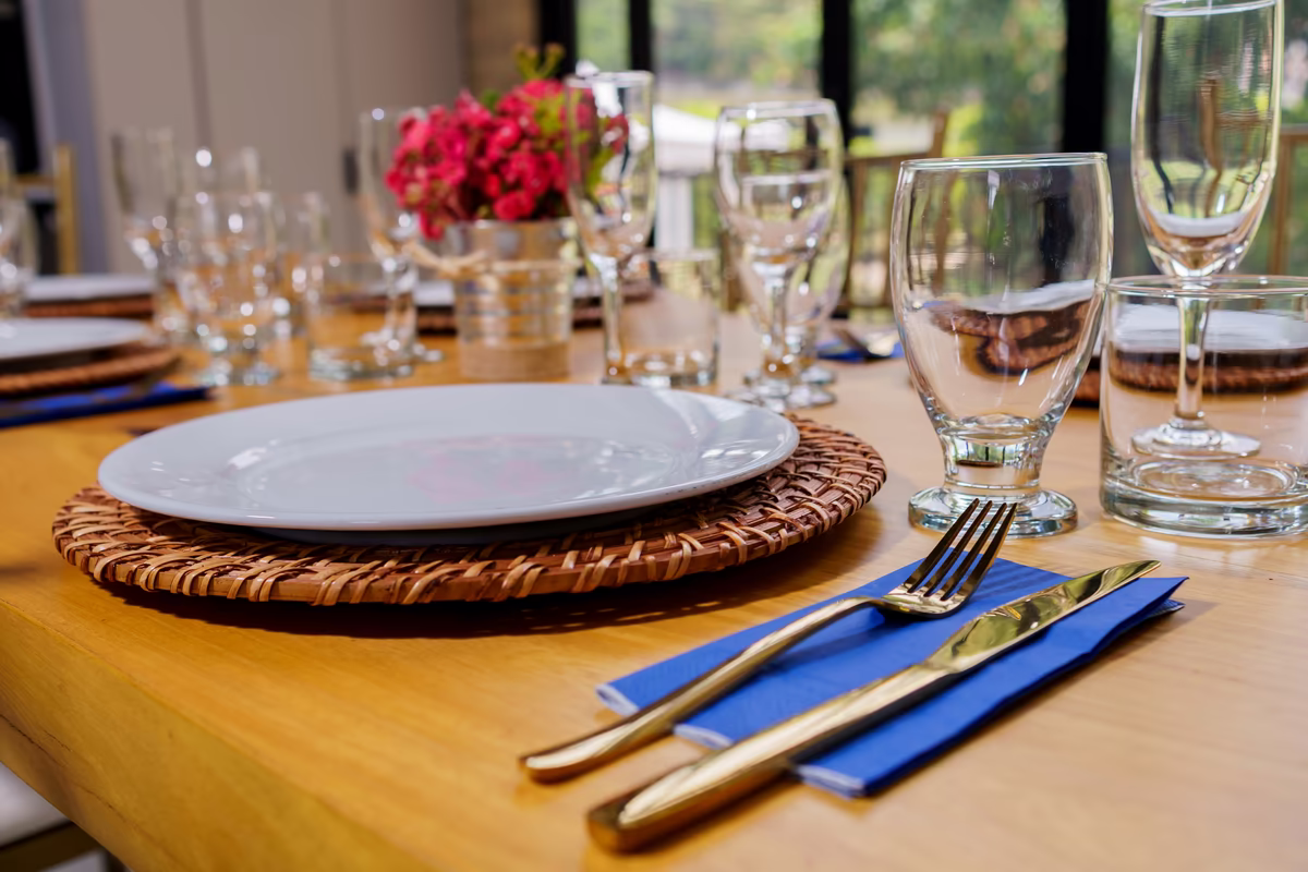 Puesto de mesa sobre madera con bajoplato tejido, servilleta azul, cubiertos dorados y copas