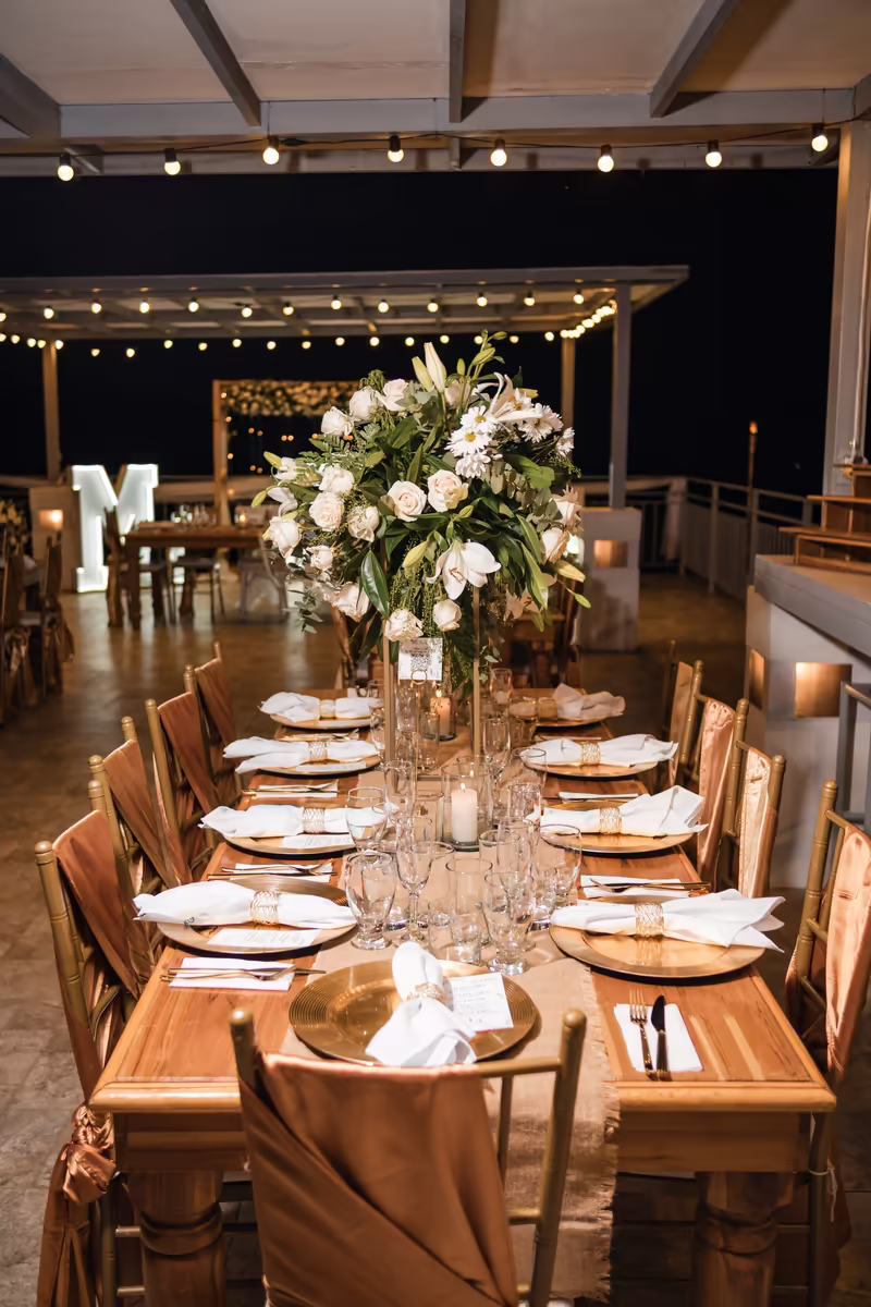 Mesa larga de recepción con centros florales blancos altos durante una boda nocturna
