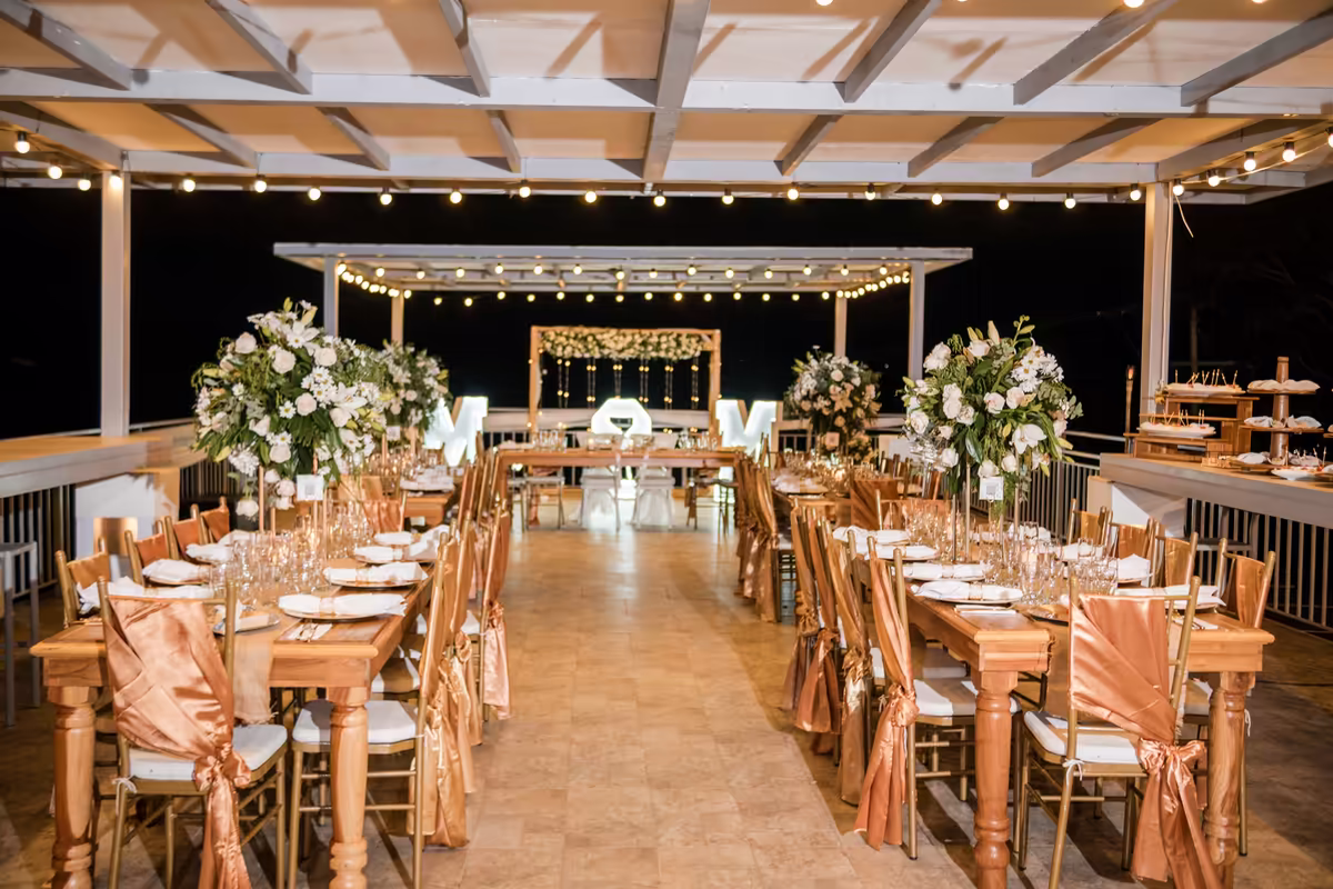 Salón de recepción montado con mesas largas y luces colgantes durante una boda nocturna