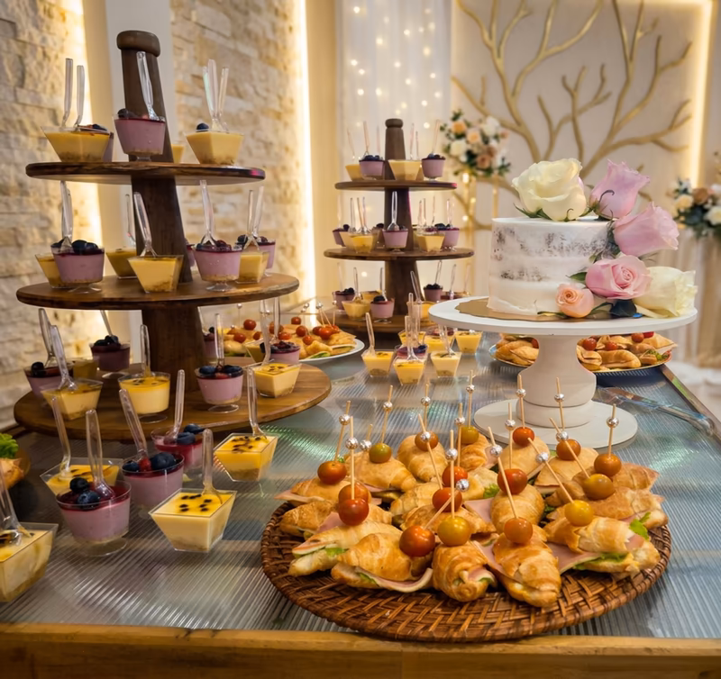 Mesa de dulces con mousses en vasitos, cupcakes, torta blanca con rosas y luces cálidas al fondo